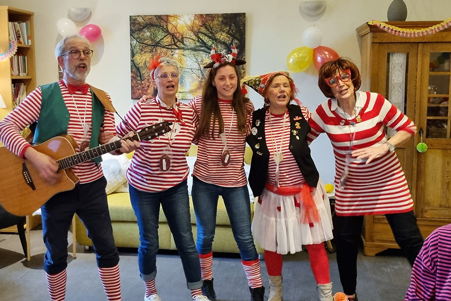 Team und Ehrenamtler vom Hospiz in Frechen e. V. Schunkeln in den Räumlichkeiten des Hospizes zu einem auf der Gitarre begleiteten Karnevalslied. Das Team ist als rot-weiße Ringelclowns verkleidet.